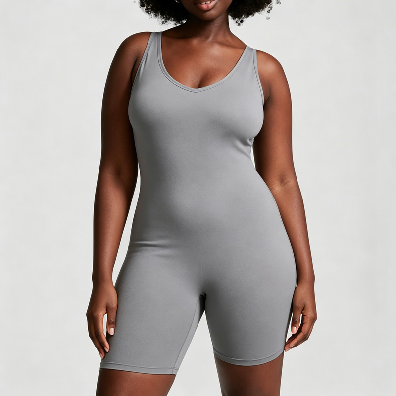 Woman standing naturally with both arms relaxed at her sides, wearing a fitted light gray sleeveless unitard shorts jumpsuit with a scoop neckline.