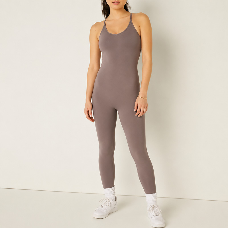 Woman standing with one leg slightly forward and both arms relaxed, wearing a taupe fitted full-length unitard with thin straps.