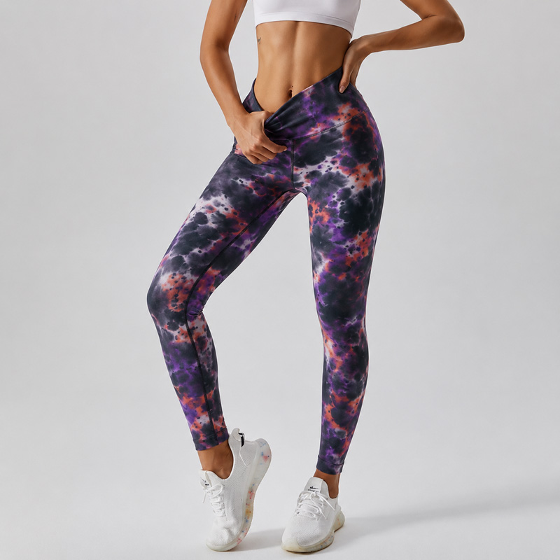 Woman standing at an angle with one hand adjusting the waistband, wearing tie-dye leggings with white sneakers.