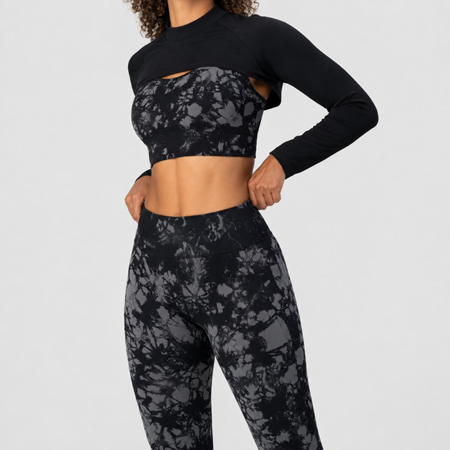 Woman standing with one hand adjusting the waistband, wearing a black marble print long sleeve crop top, bra, and matching leggings.