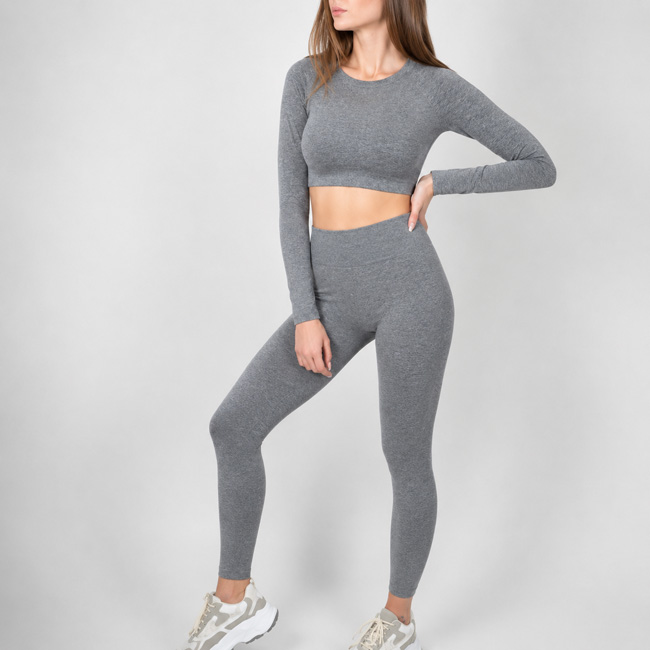 Woman posing with one hand on her waist and one leg slightly bent, wearing a gray long sleeve crop top and fitted leggings set.