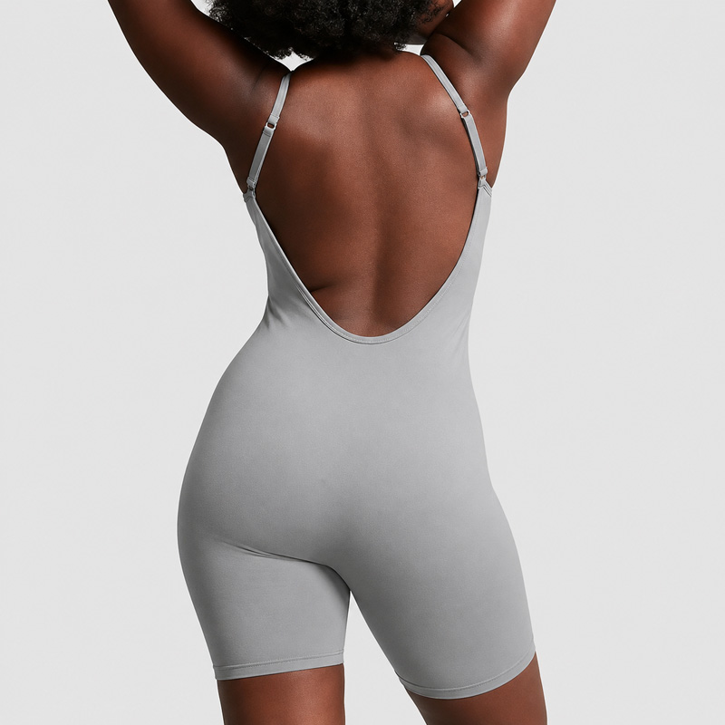 Back view of a woman raising both arms above her head while wearing a light gray backless sleeveless unitard bodysuit with thin straps and fitted shorts.
