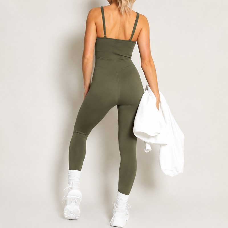 Back view of a woman walking forward while holding a white shirt in one hand, wearing an olive green strappy full-length jumpsuit.