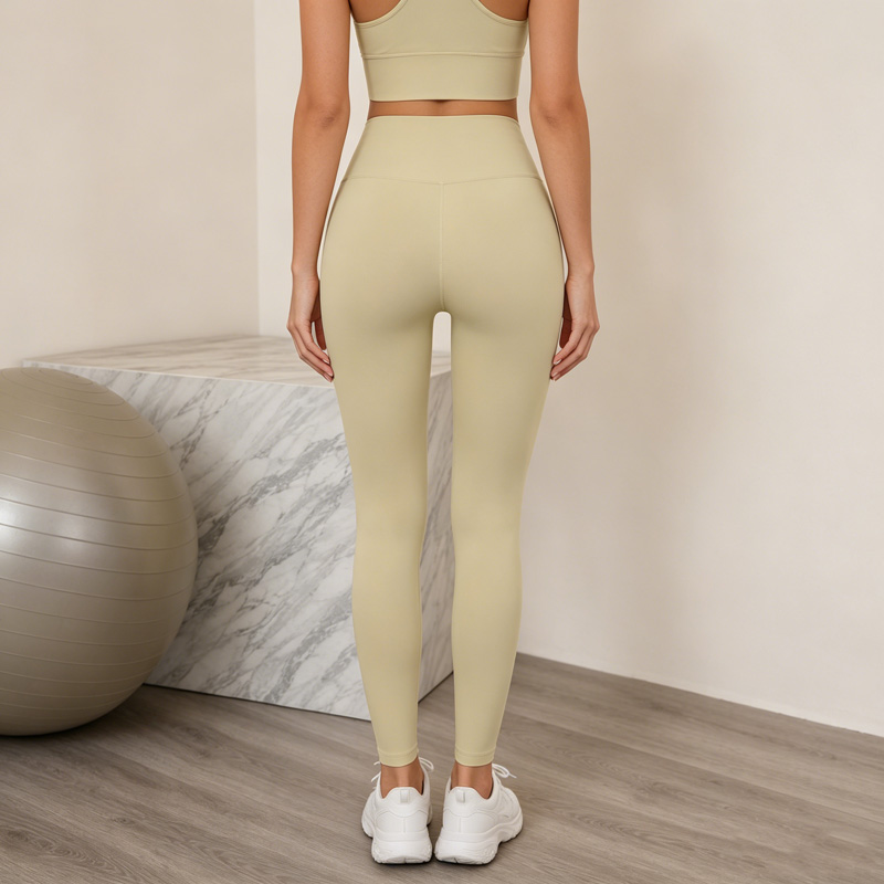 Back view of a woman standing naturally while wearing beige ankle-length leggings paired with white sneakers.